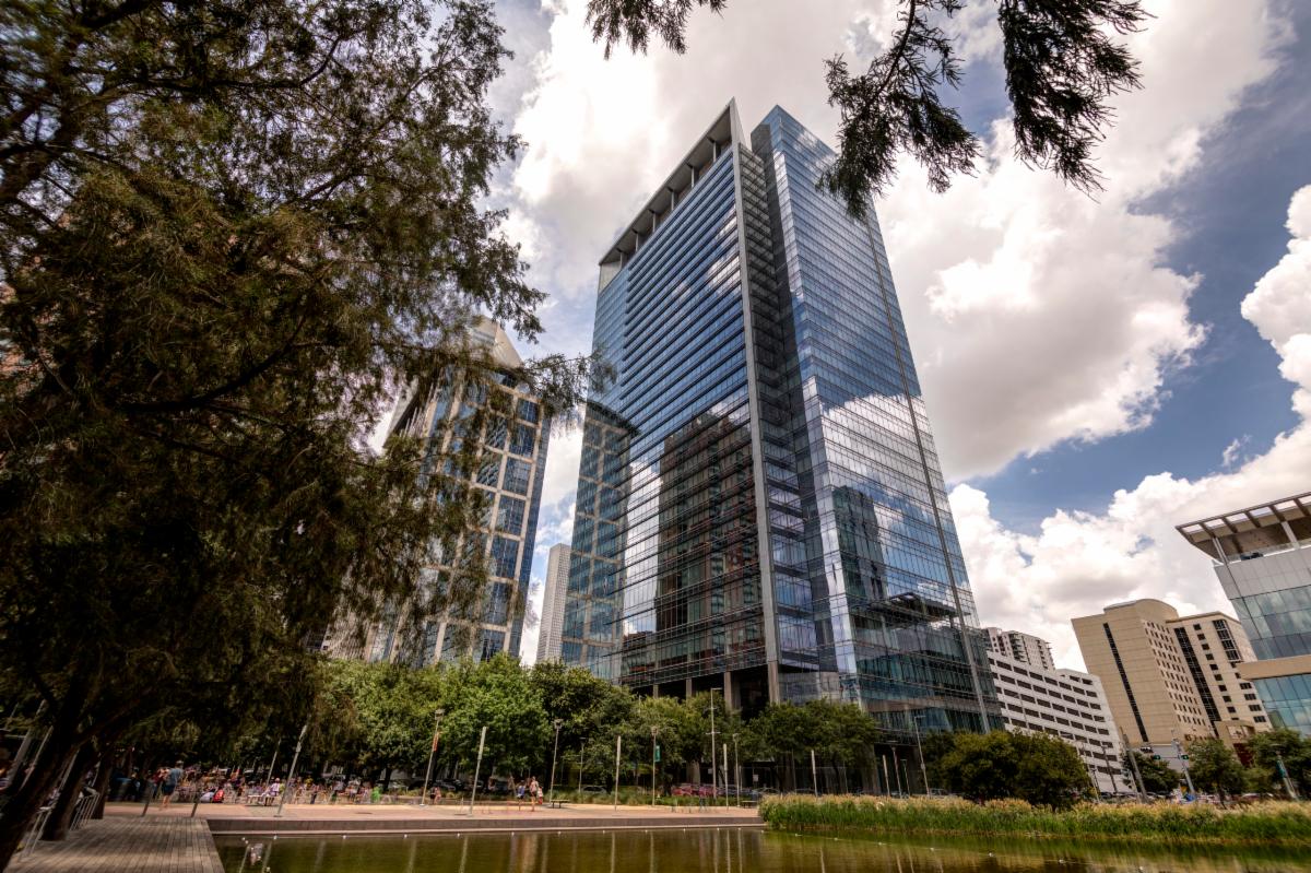 A modern glass skyscraper reflecting the sky and surrounding trees.