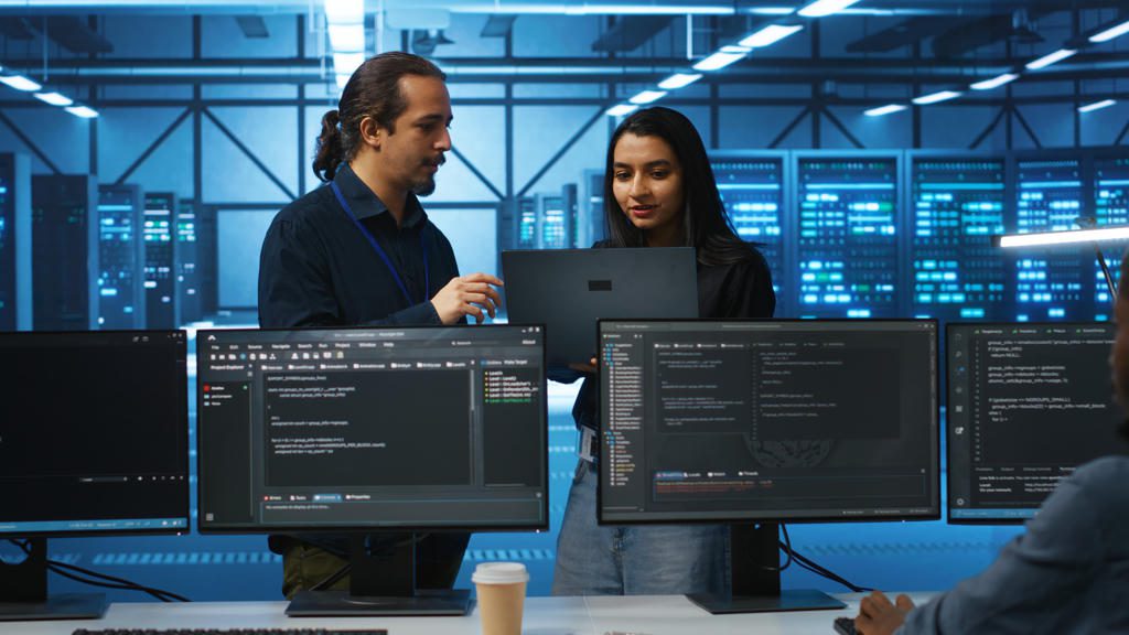Two software developers collaborate in a modern tech workspace with code on multiple screens.